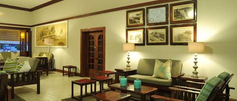 A cozy living room with a sofa, armchairs, and a coffee table. There are framed pictures on the wall and a door leading to another room.