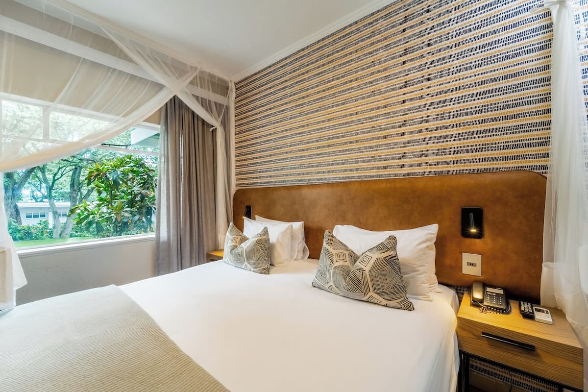 A hotel room with a large bed, two patterned pillows, a nightstand with a phone, and a view of greenery outside.