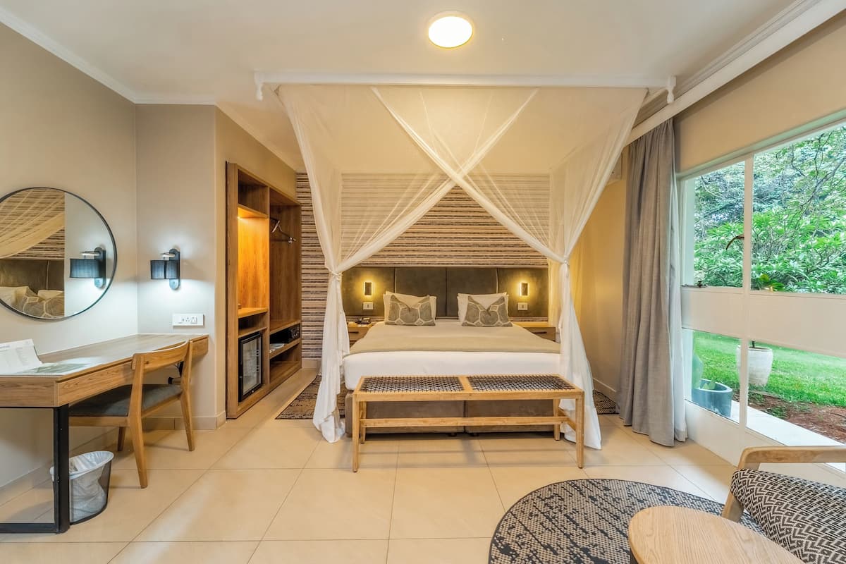 A bedroom with a canopy bed, a desk with a chair, a round table, and a large window with a view of greenery.