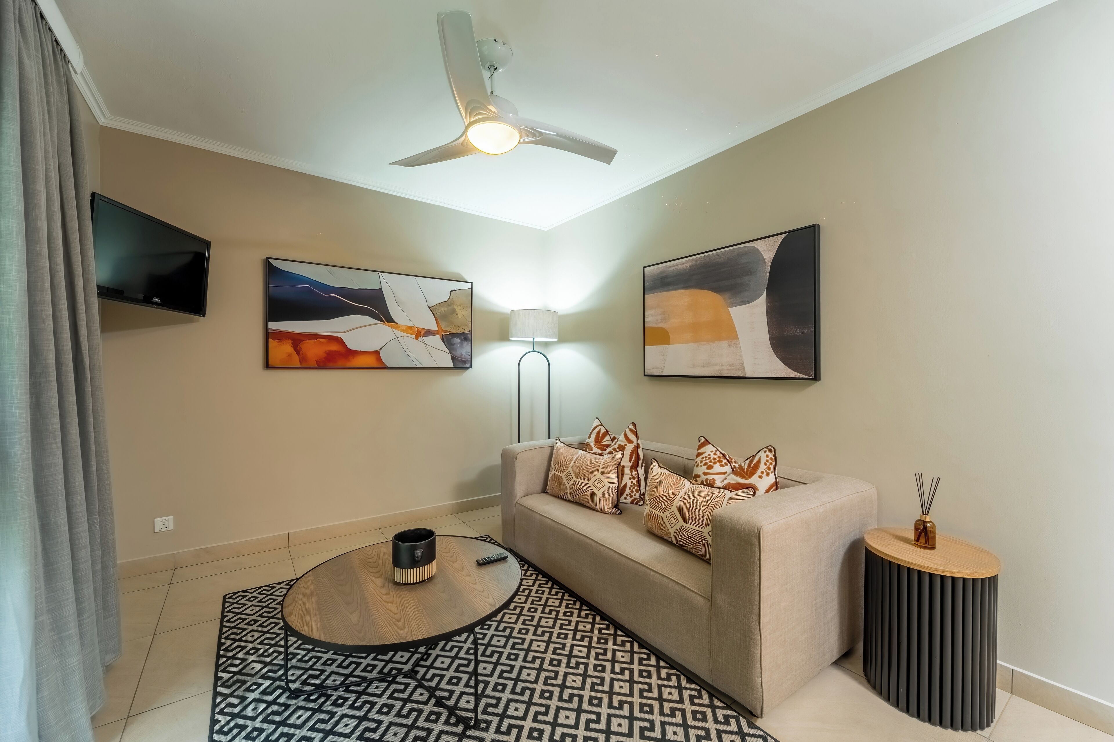 Un salon comprenant un canapé, une table basse, un téléviseur, des œuvres d’art abstraites accrochées au mur, un ventilateur de plafond et une lampe.