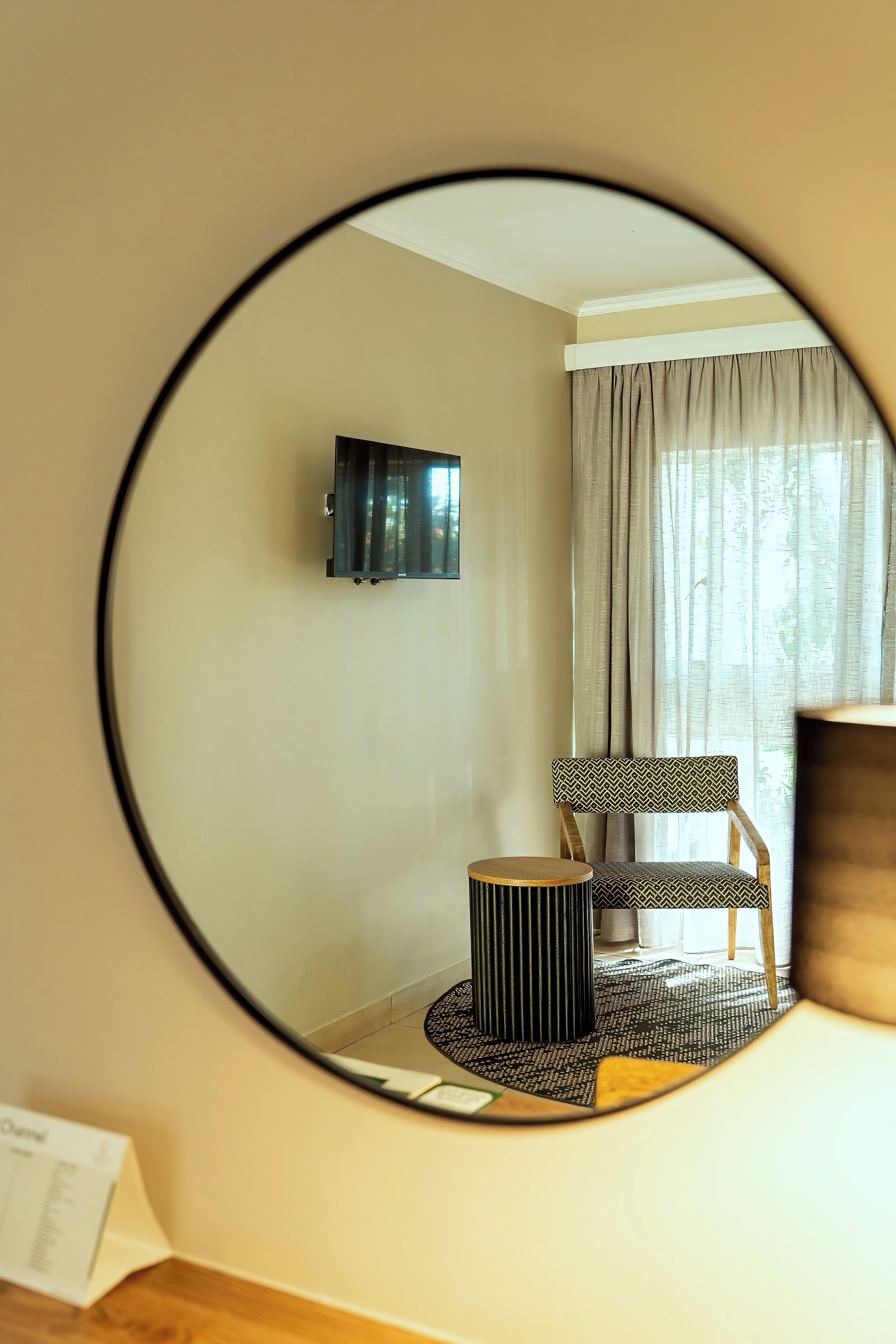 A room with a round mirror reflecting a chair, a small table, and a television on the wall.