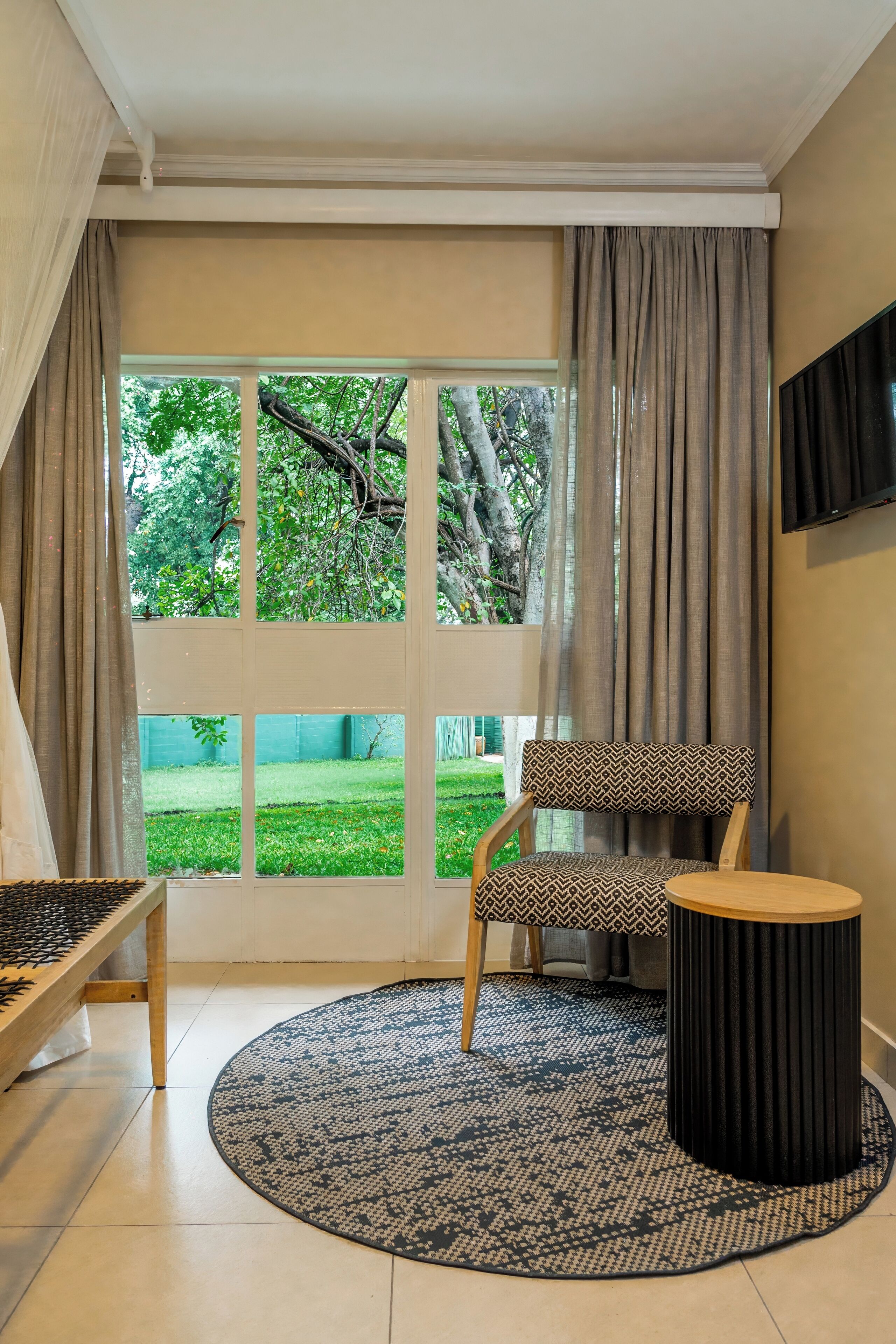 Una habitación con un ventanal grande, un sillón estampado, una mesa redonda y un reposapiés negro.