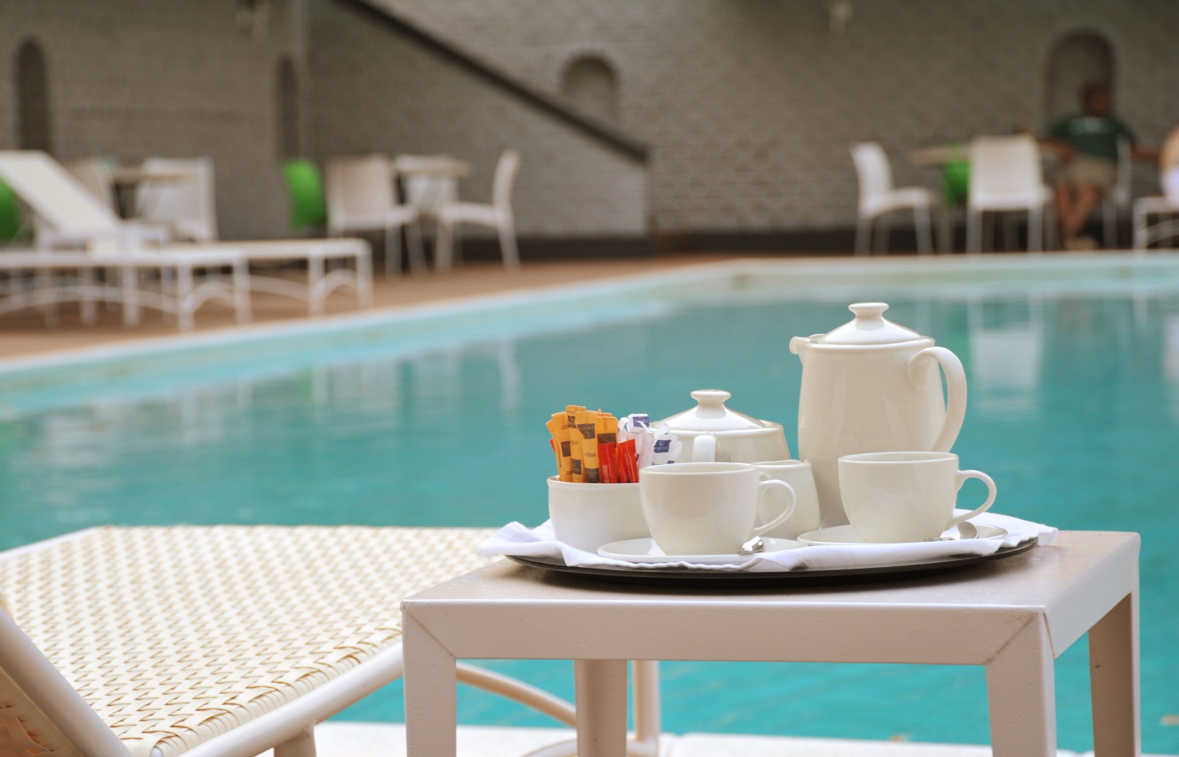 A poolside table with a tray of white teacups, a teapot, and a sugar packet holder.