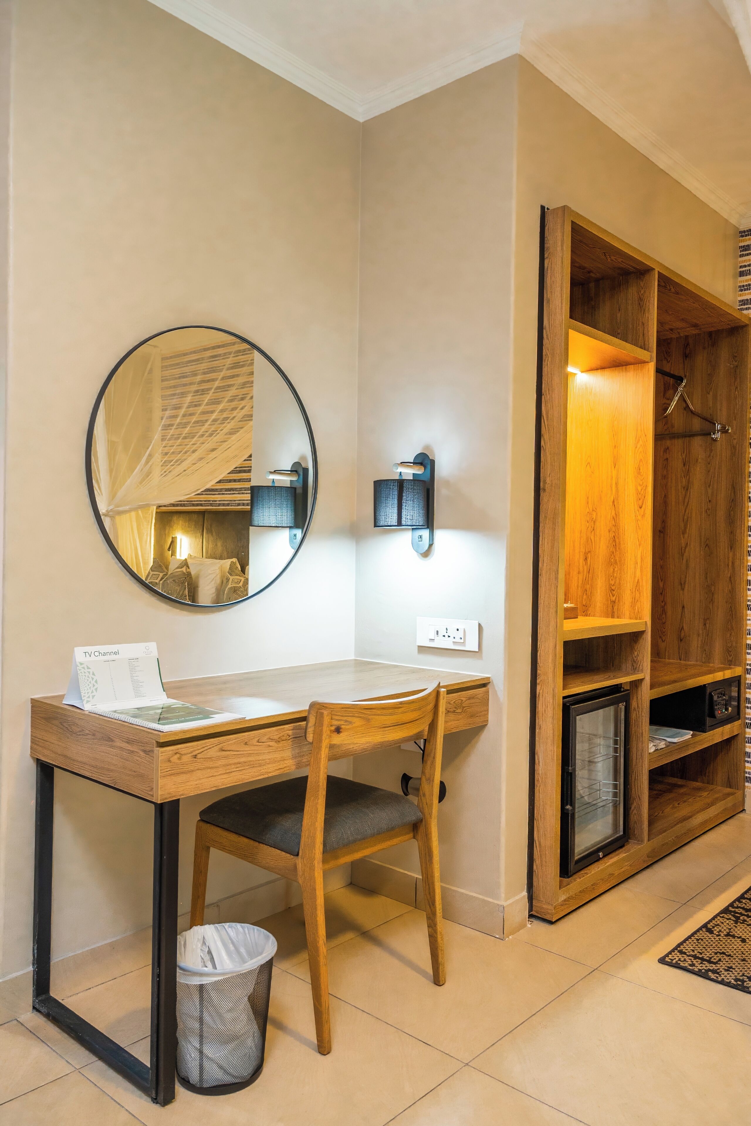 A hotel room with a wooden desk, a chair, a round mirror, a TV channel brochure, a trash bin, and a wooden wardrobe with a hanging rack.