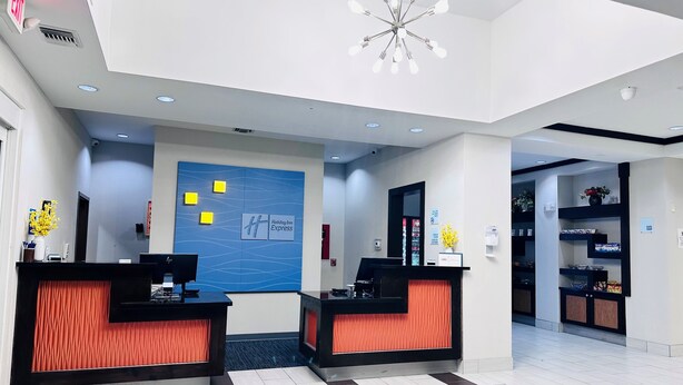 Ein Hotel-Empfangsbereich mit zwei Empfangstresen, einer blauen Wand mit dem Holiday Inn Express Logo, einer modernen Pendelleuchte und einem kleinen Geschäft im Hintergrund.