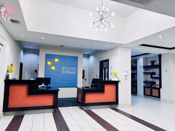 A hotel reception area with two reception desks, a blue wall with the Holiday Inn Express logo, a modern chandelier, and a small shop in the background. - Holiday Inn Express & Suites Houston East - Baytown by IHG (Baytown)