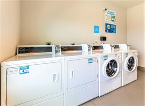 A laundry facility with four washing machines and a dryer, a control panel, and a poster on the wall. - Holiday Inn Express & Suites Houston East - Baytown by IHG (Baytown)
