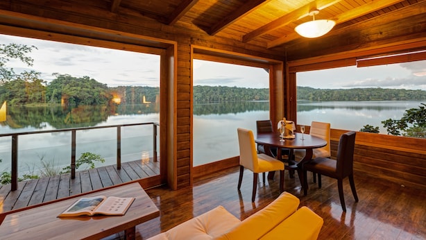 Une terrasse en bois avec une table et des chaises, donnant sur un lac paisible et des collines boisées.