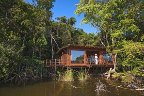 Une cabane en bois sur pilotis, entourée d’une forêt dense et bordée par un plan d’eau.