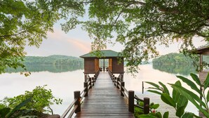 Une jetée en bois mène à un chalet doté d’un toit vert, entouré d’une végétation luxuriante et bordé d’un lac paisible.