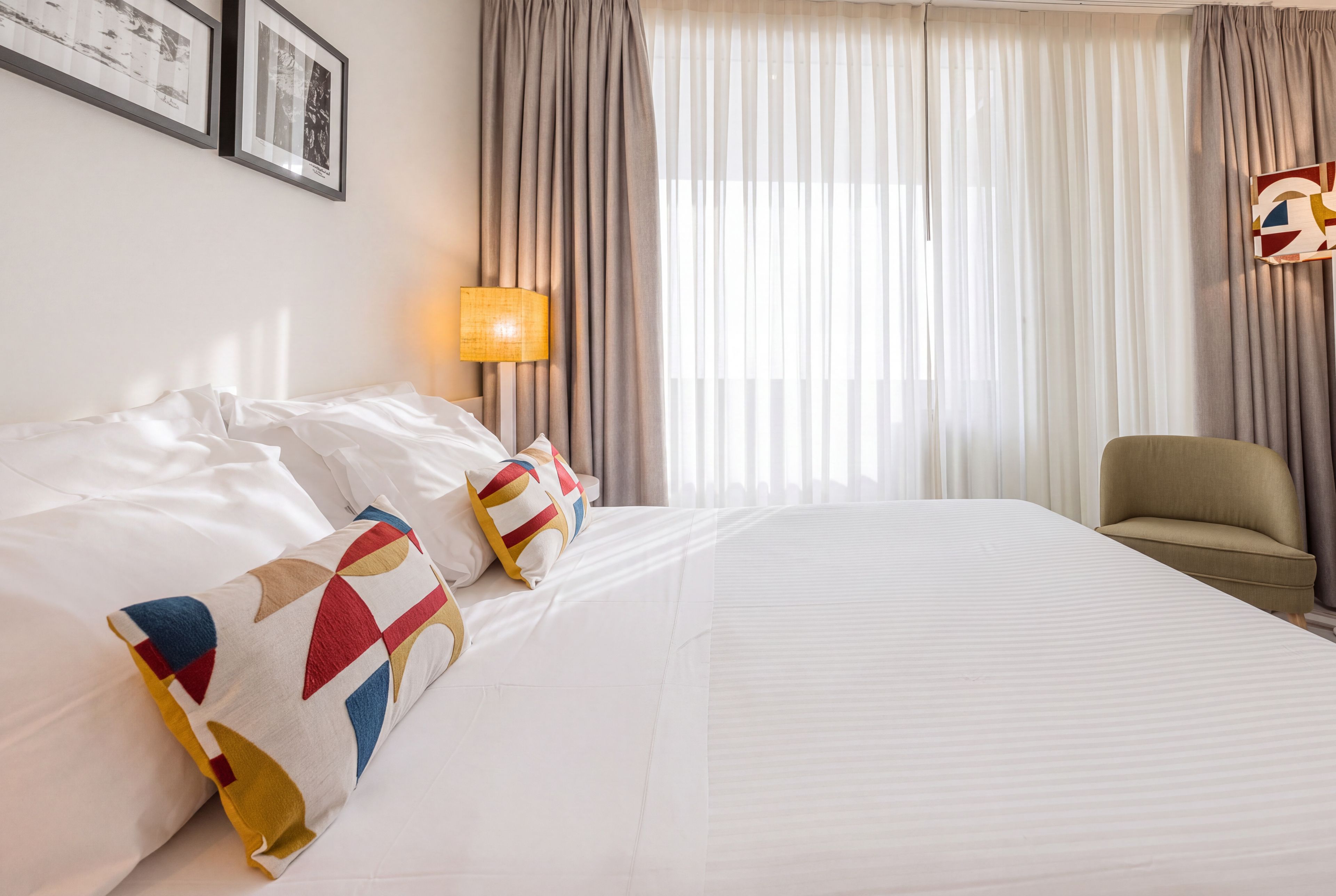 Une chambre d’hôtel avec un grand lit, deux oreillers, une table de chevet avec une lampe, une chaise et des rideaux.
