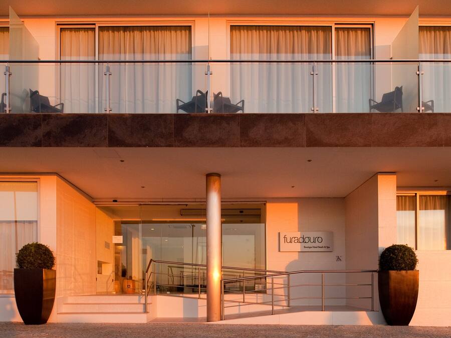 Una entrada de hotel con fachada de vidrio, una escalera que lleva al piso superior y dos plantas en macetas.