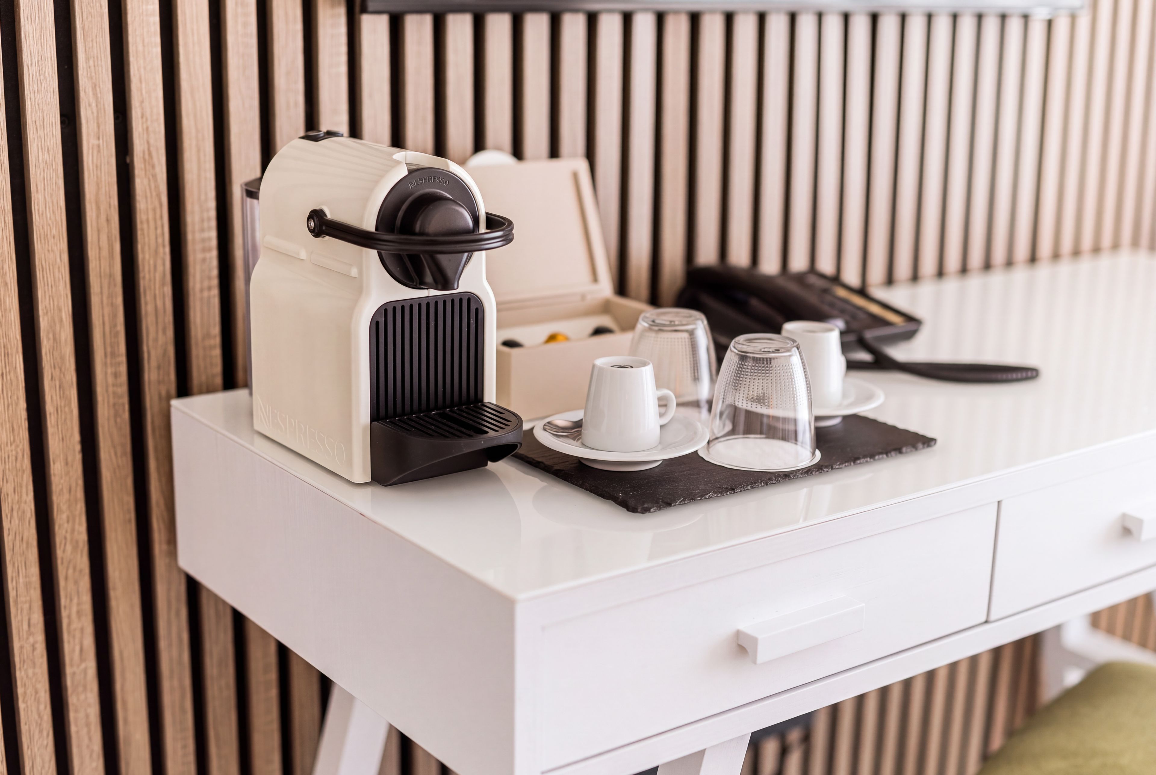 Eine weiße Kaffeemaschine mit schwarzem Fuß, eine weiße Tasse, ein Glas und eine kleine Schachtel auf einem weißen Tisch vor einer Wand aus Holzlatten.