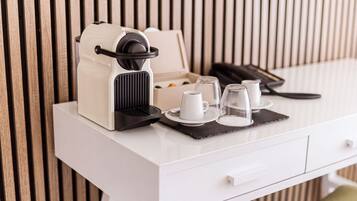 Eine weiße Kaffeemaschine mit schwarzem Fuß, eine weiße Tasse, ein Glas und eine kleine Schachtel auf einem weißen Tisch vor einer Wand aus Holzlatten.