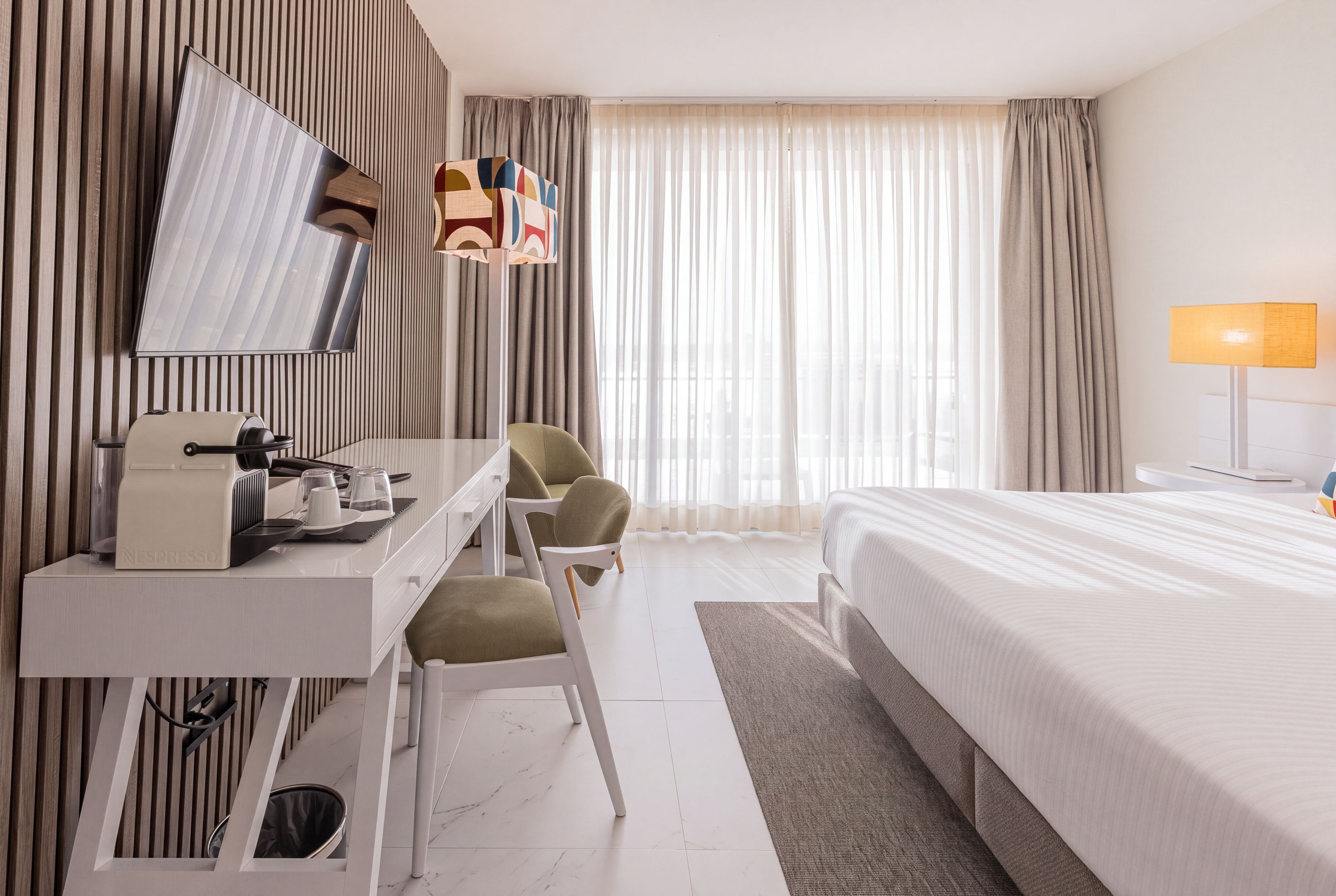 Habitación de hotel con cama, escritorio con cafetera, silla, televisor montado en la pared y ventana con cortinas.