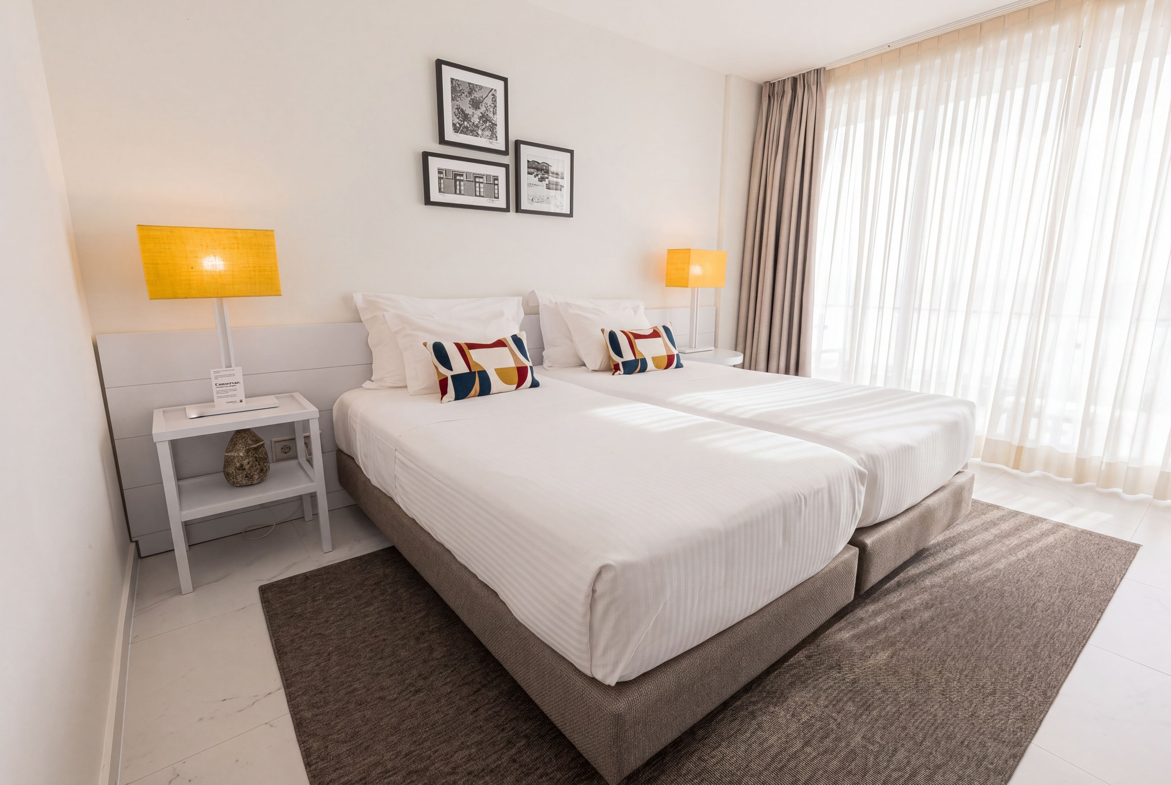 A hotel room with two beds, a nightstand, a lamp, a rug, and a window with curtains.