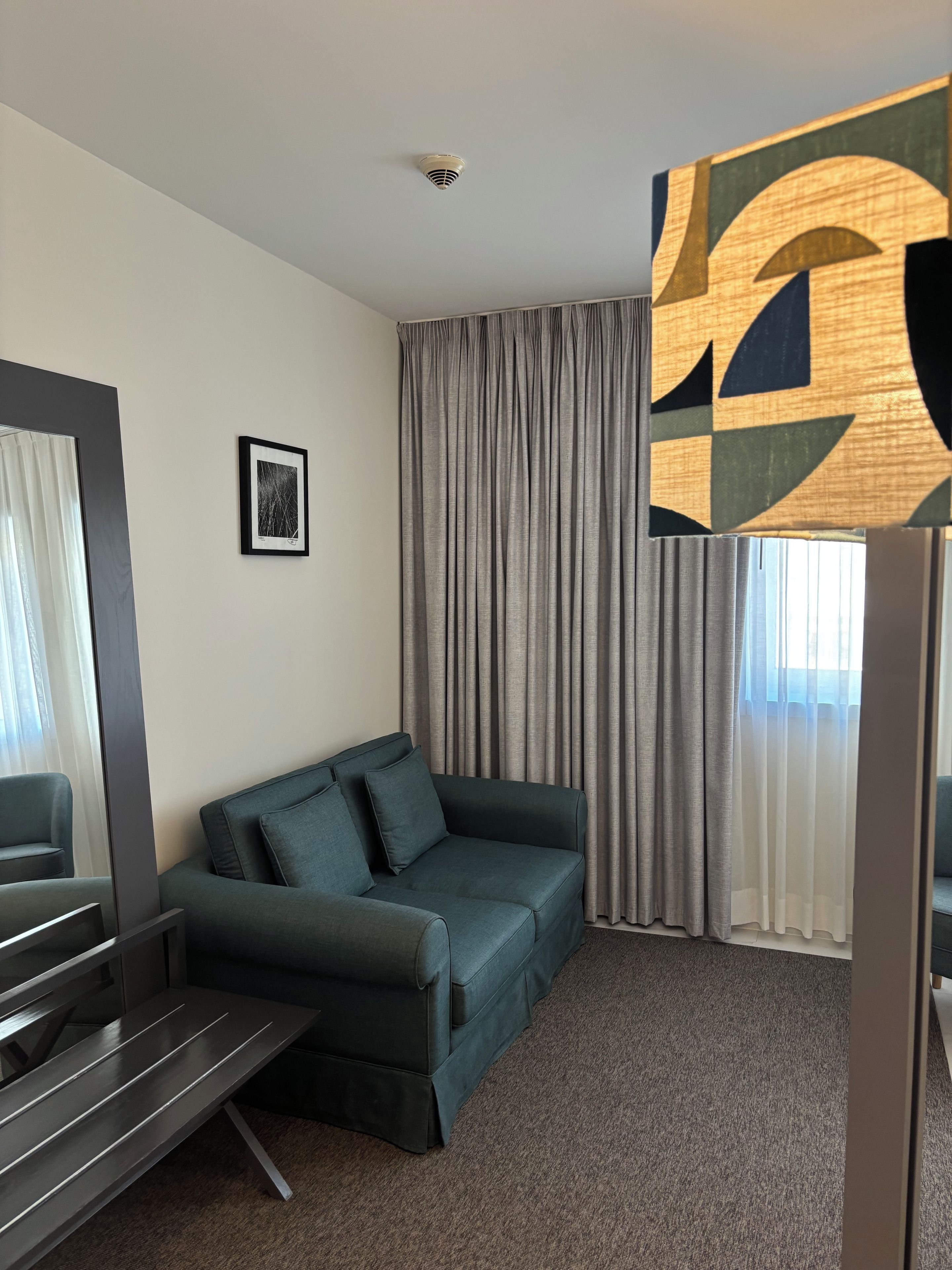 Quarto de hotel moderno com um sofá verde escuro, uma moldura na parede e uma grande obra de arte abstrata sobre a janela.