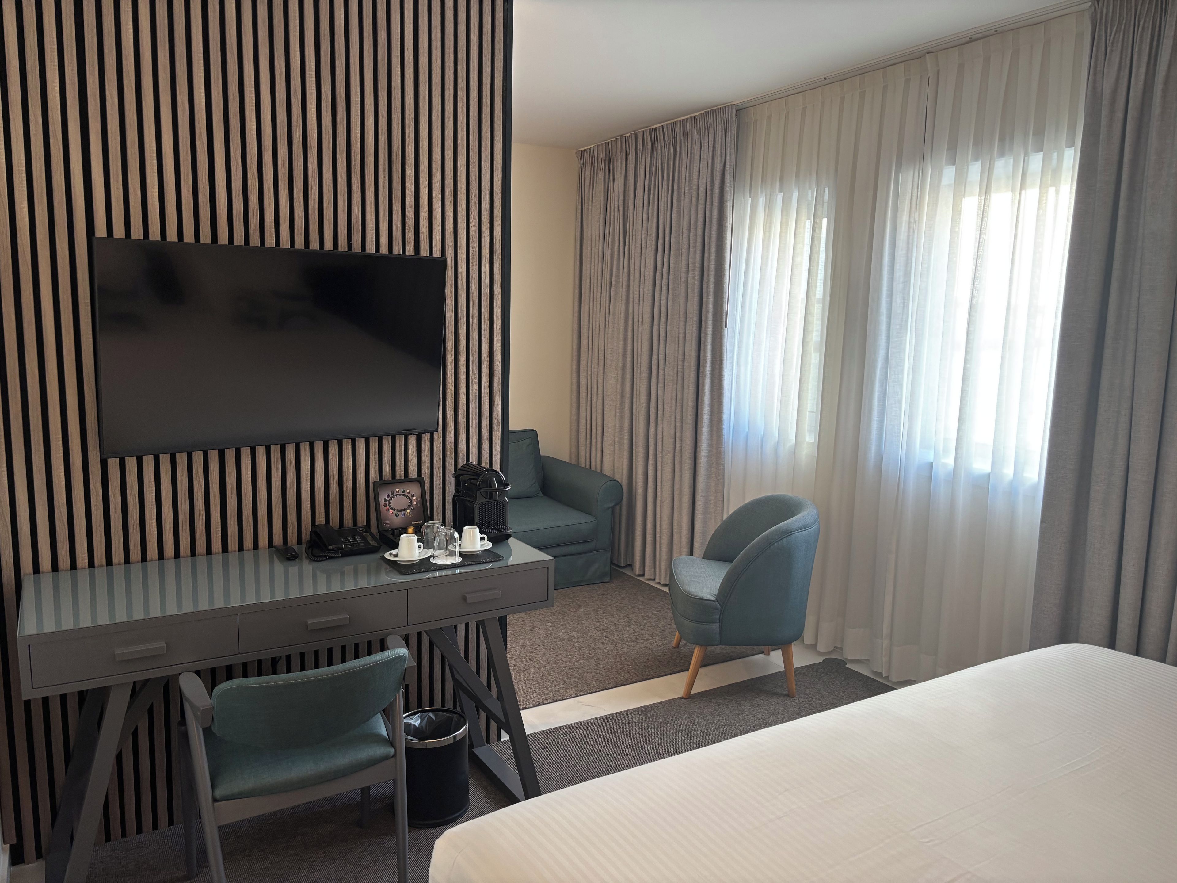 A hotel room with a flat-screen TV mounted on a wall, a desk with a chair, a sofa, a bed, and a window with curtains.