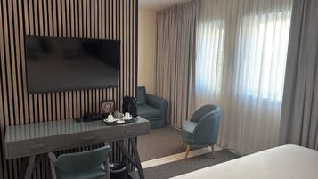 A hotel room with a flat-screen TV mounted on a wall, a desk with a chair, a sofa, a bed, and a window with curtains.