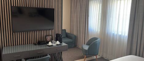 A hotel room with a flat-screen TV mounted on a wall, a desk with a chair, a sofa, a bed, and a window with curtains.