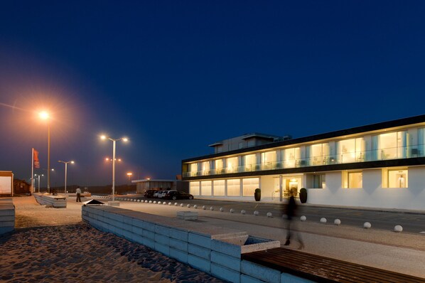 Un bâtiment moderne doté d’un toit plat et de grandes fenêtres, situé près d’une zone sablonneuse, avec une promenade dallée et des lampadaires.