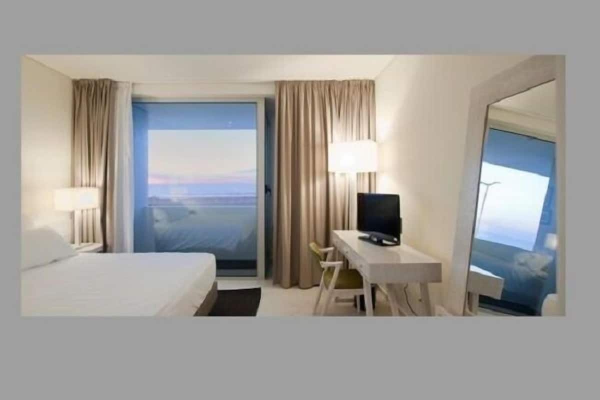 Habitación de hotel con cama, escritorio con computadora, silla, televisor y una ventana grande con vistas.