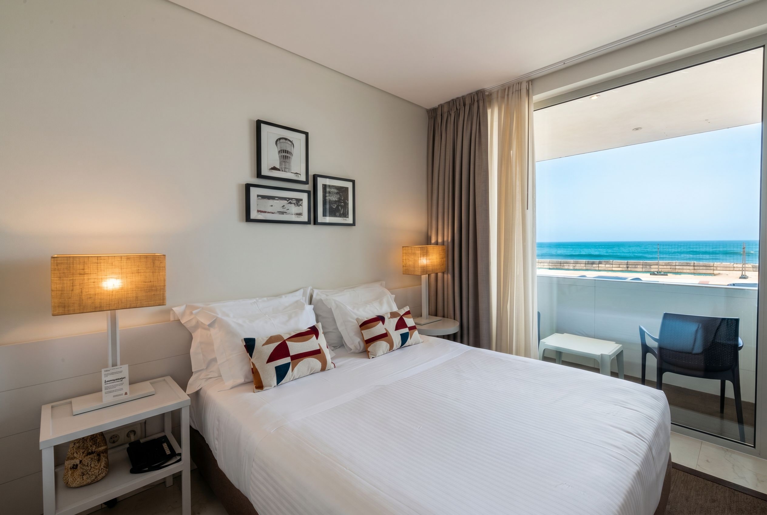 Een hotelkamer met een bed, twee bedlampen, een klein tafeltje, een stoel en een balkon met uitzicht op zee.