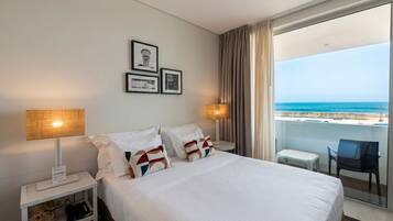 Een hotelkamer met een bed, twee bedlampen, een klein tafeltje, een stoel en een balkon met uitzicht op zee.