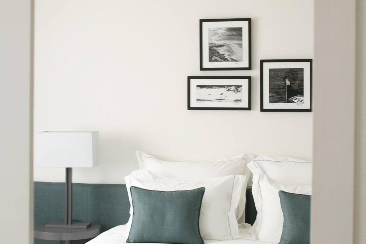 Una cama bien hecha con almohadas blancas y azul turquesa, una mesita de noche con lámpara y tres cuadros en la pared.