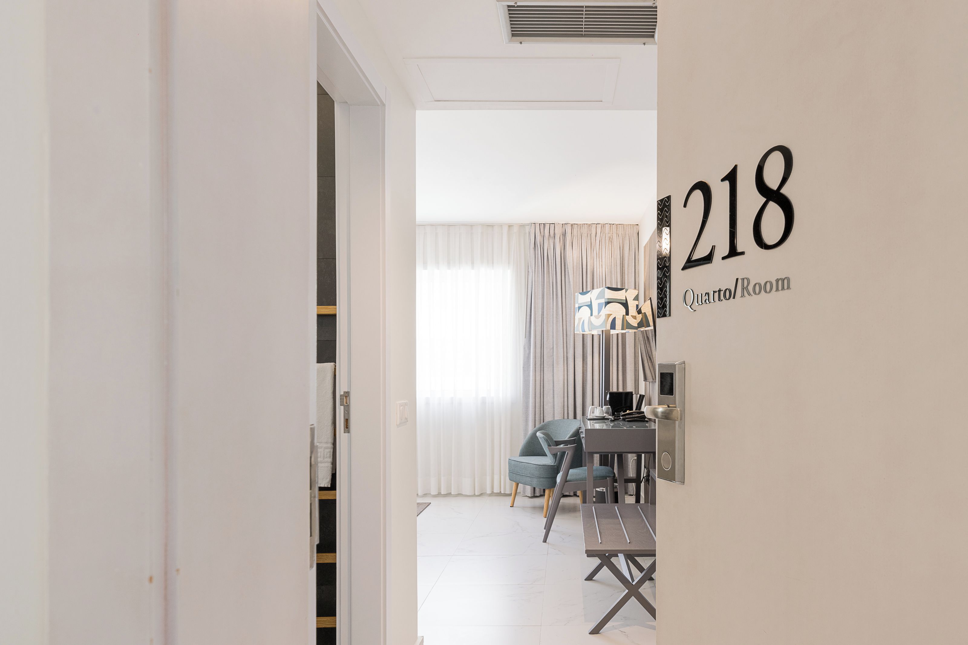 A hotel room with a number 218 sign, a dining area with a table and chairs, and a large window with curtains.