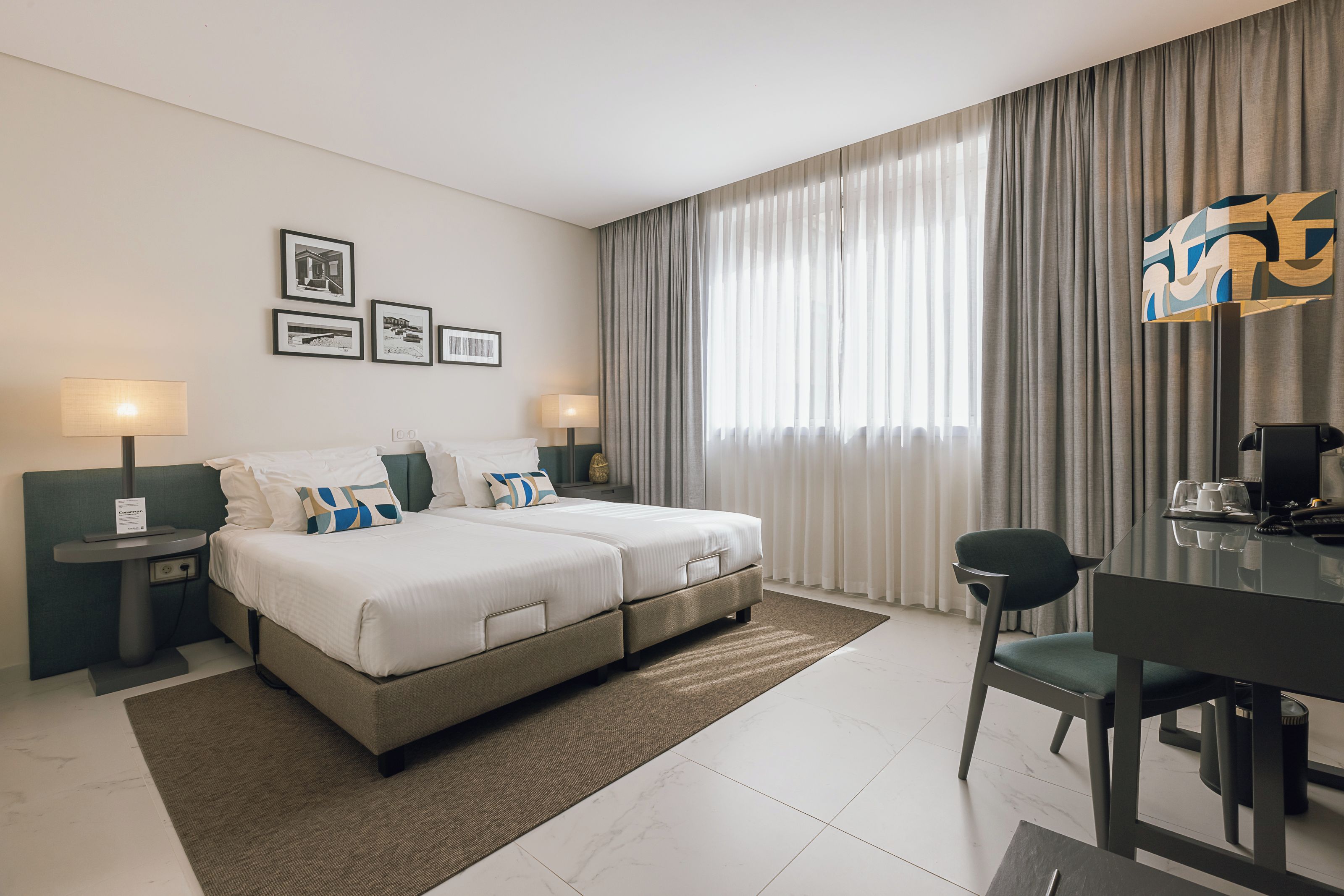 A hotel room with a bed, bedside tables, a desk, and a chair. There are curtains, a window, and framed pictures on the wall.