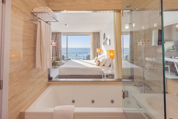 Habitación de hotel moderna con ducha envuelta en vidrio, cama grande con ropa blanca, vista al mar y balcón con mobiliario exterior.