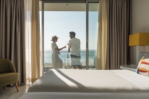 A hotel room with a large bed, a view of the sea, and a balcony with curtains.