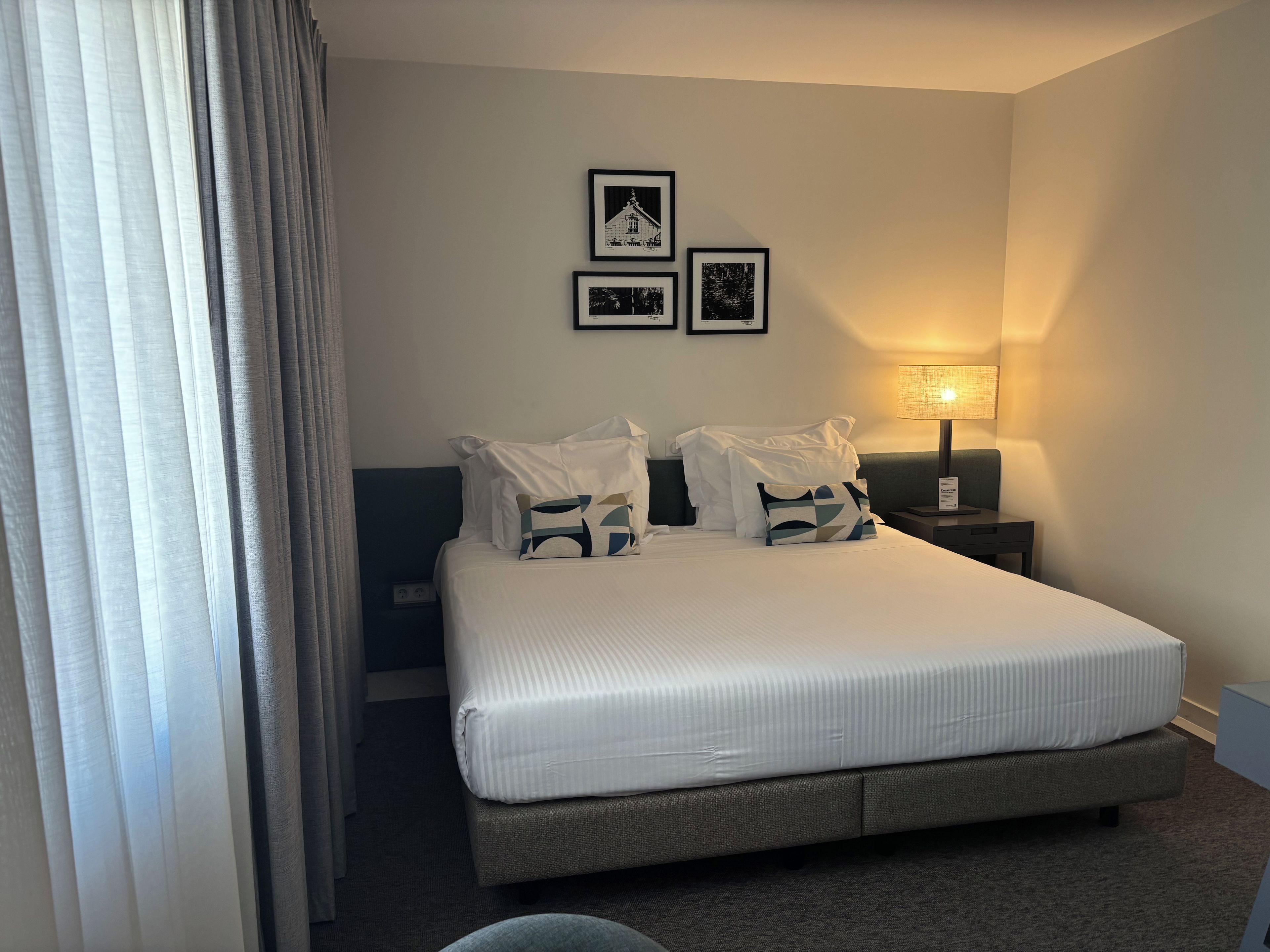Habitación de hotel con una cama grande, dos mesitas de noche, una lámpara y un ventanal con cortinas.