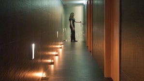 Un pasillo iluminado por pequeñas velas en el suelo, con una persona caminando hacia una puerta.