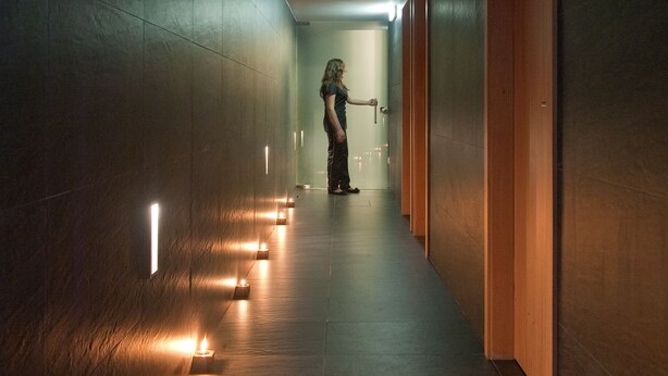 Un couloir dans lequel une personne marche en direction d’une porte, éclairé par de petites bougies posées sur le sol.