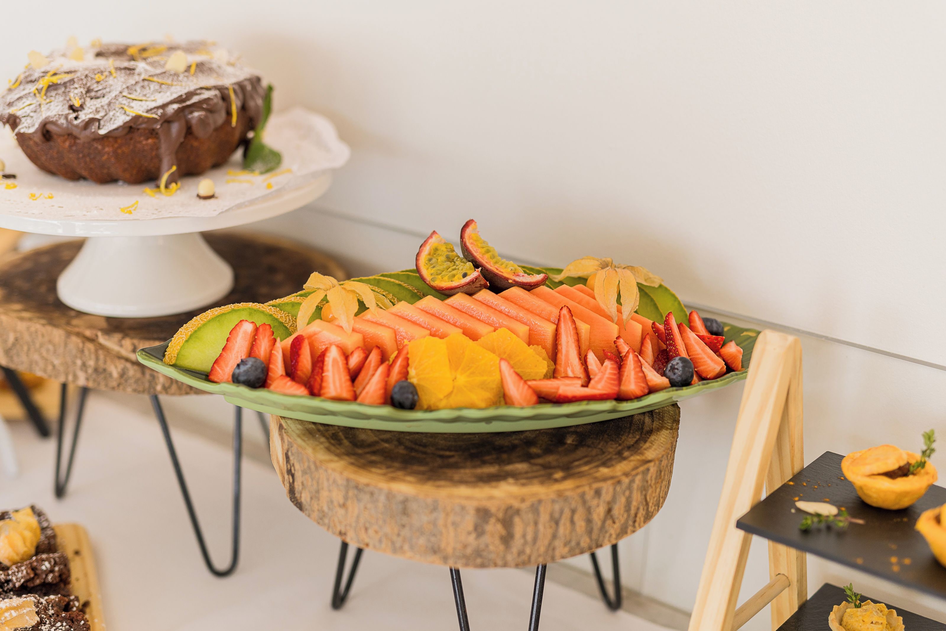 A fruit platter on a wooden stand with a loaf of bread on a white cake stand in the background.