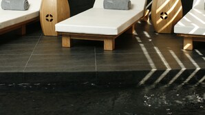 Tres tumbonas con toallas, taburetes de madera y una pared de baldosas negras con listones de madera.