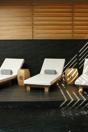 Trois chaises longues avec des serviettes, des tabourets en bois et un mur recouvert de carreaux noirs avec des lattes de bois.