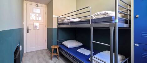 A dormitory room with bunk beds, a small stool, a door with a bulletin board, and blue lockers.