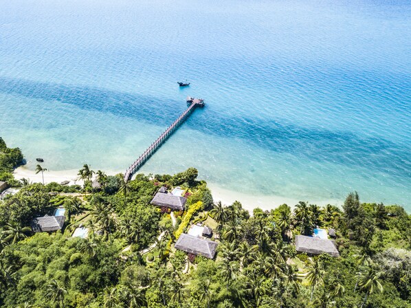 An aerial view of a tropical resort with a long pier extending into clear blue waters, surrounded by lush greenery and thatched-roof huts. - Zanzi Resort (Kama)