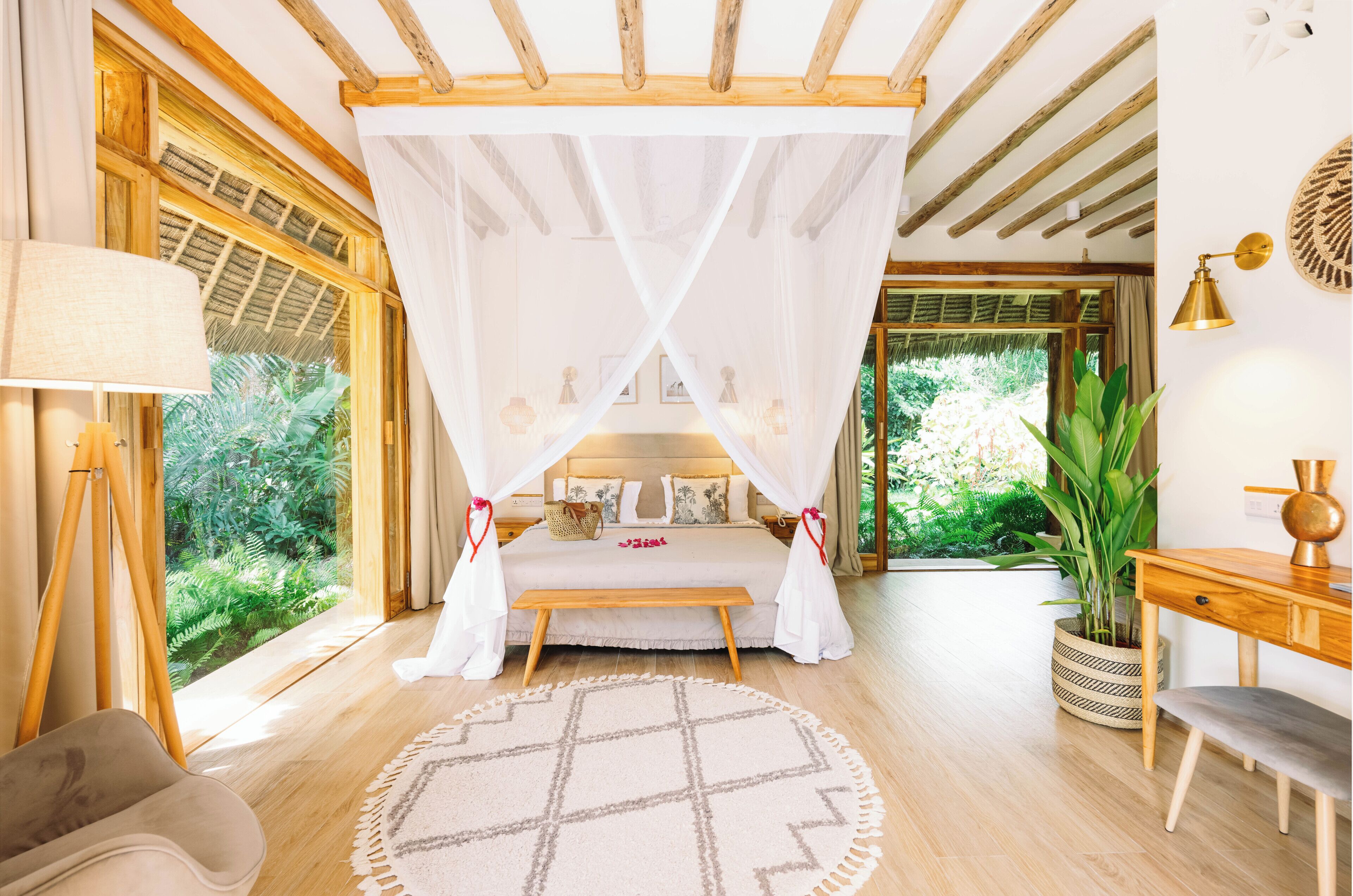 A bedroom with a canopy bed, a bedside table, a lamp, a chair, a desk, and a view of greenery.