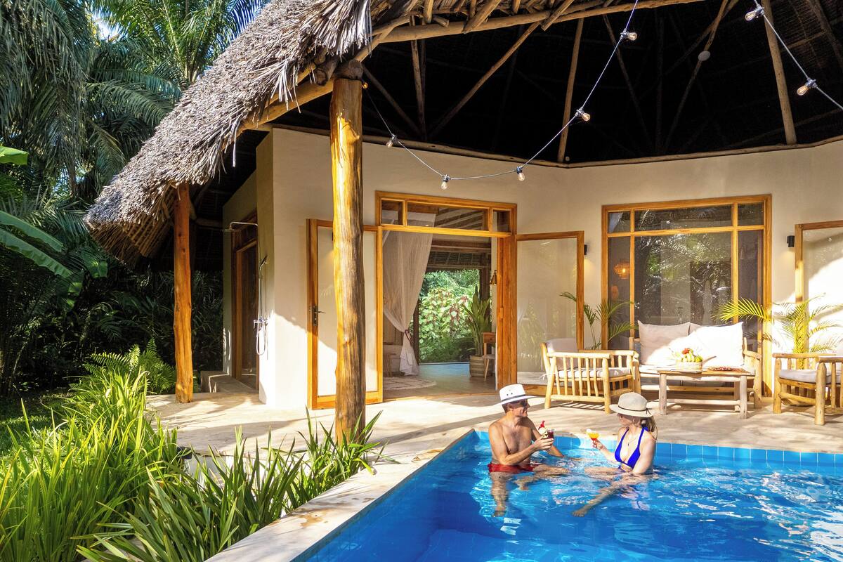Un área junto a la piscina con techo de paja, mobiliario de madera y vistas a exuberante vegetación.