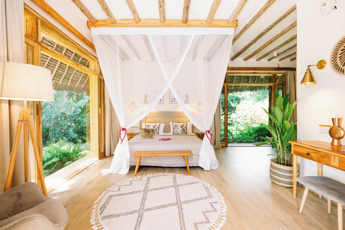 A bedroom with a canopy bed, wooden flooring, a round rug, a desk, and a potted plant.
