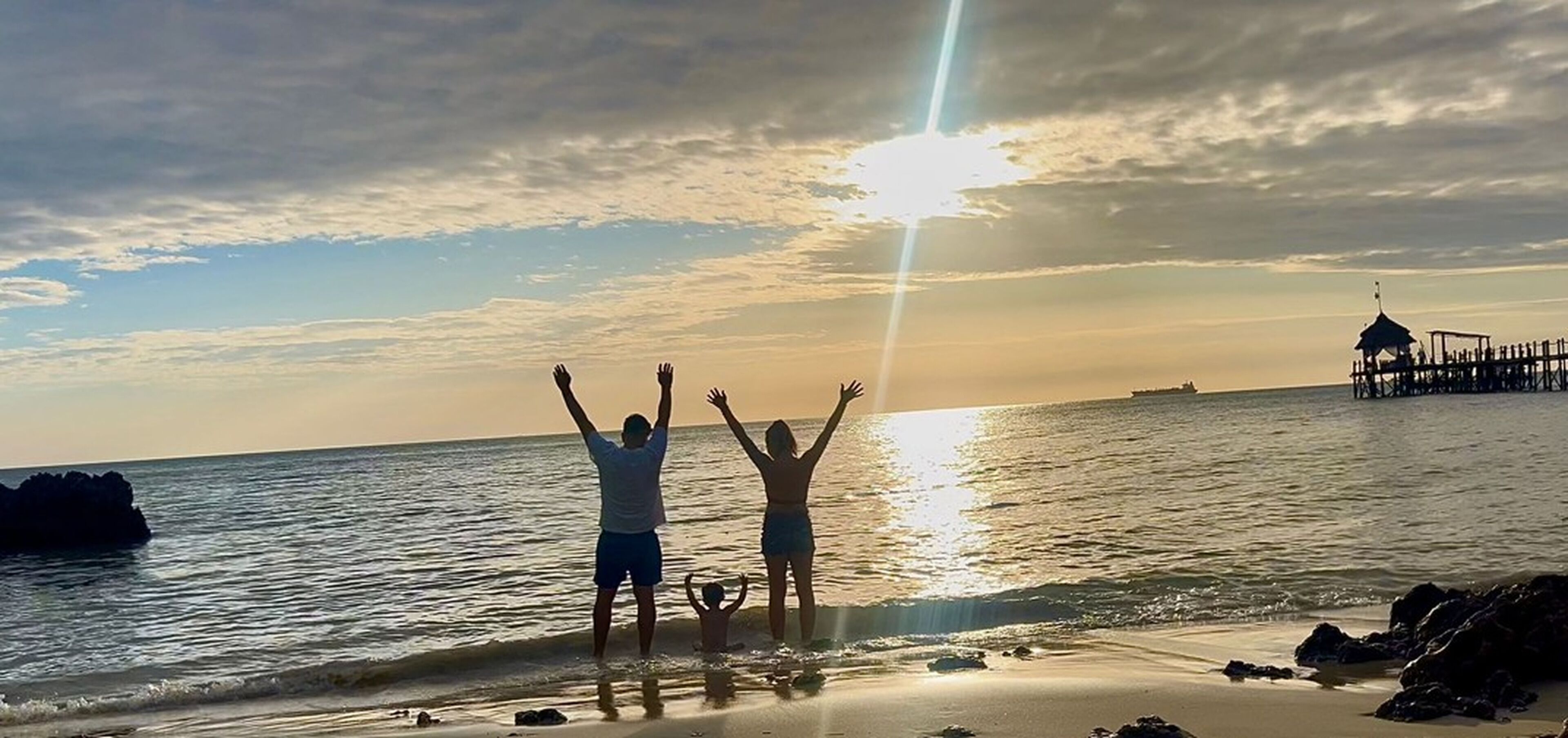 Escena de playa con un atardecer, un muelle y una persona alzando las manos.