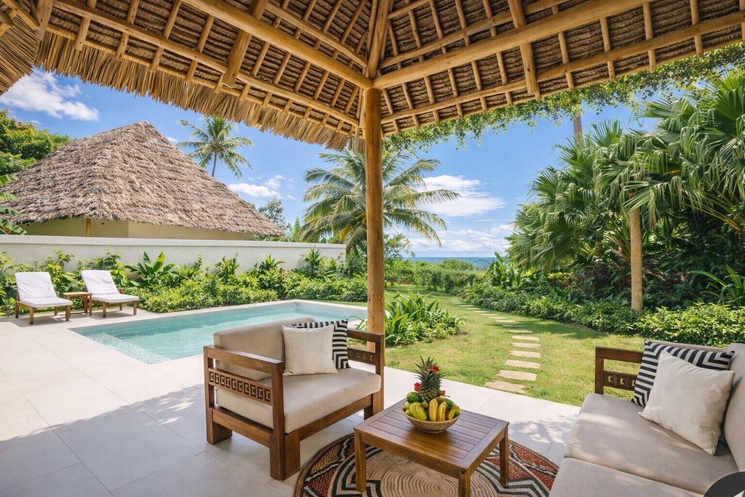 Zona de piscina con techo de paja, sillas de descanso, una mesa de madera con frutero y vistas a un jardín exuberante y al océano.