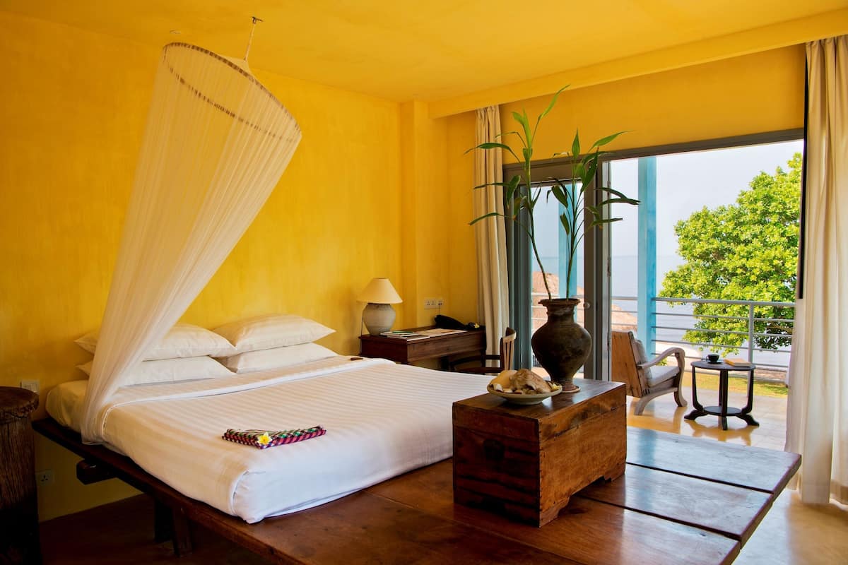A room with a bed, bedside table, lamp, and a view of greenery outside.