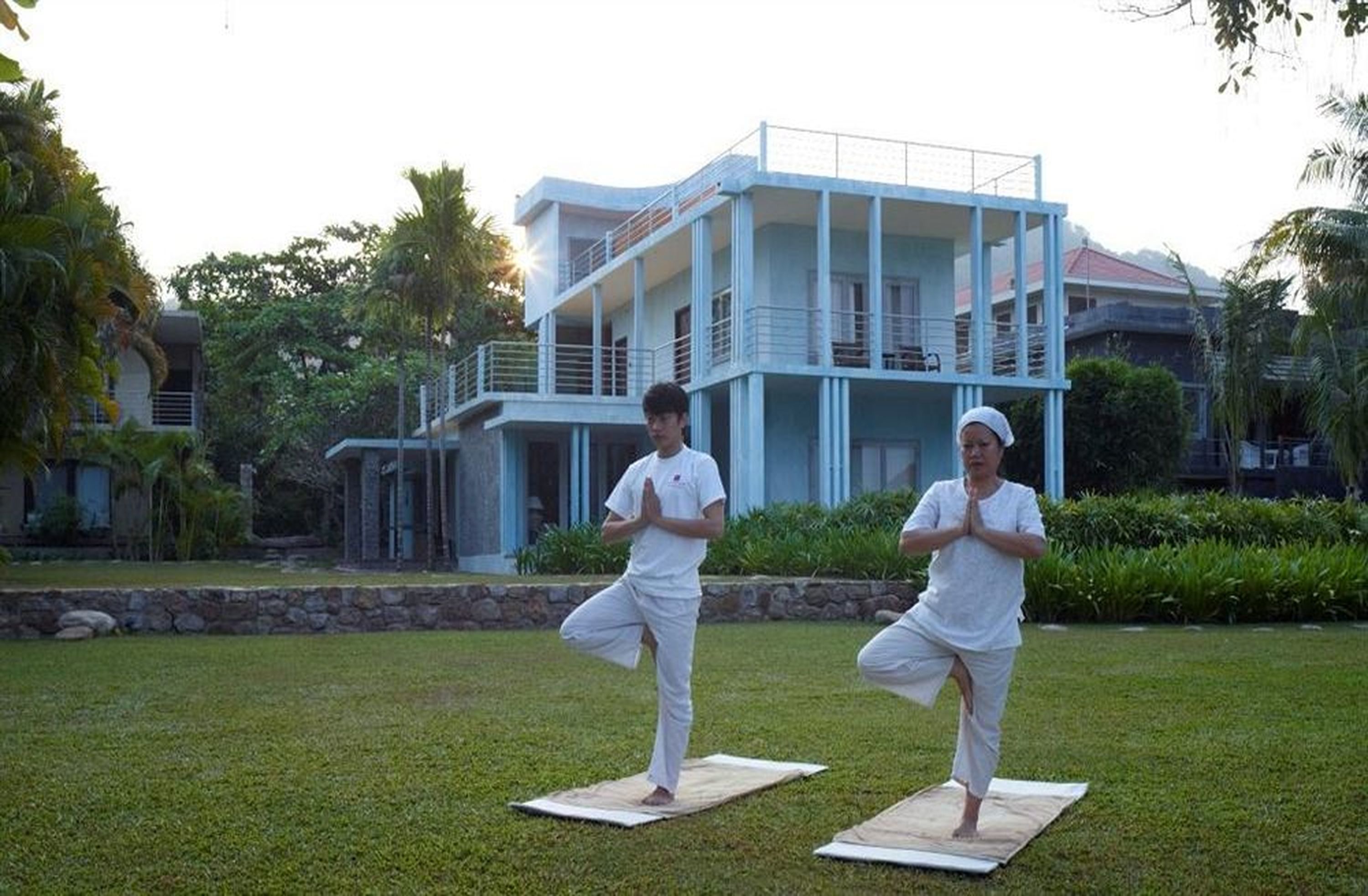 Twee personen doen yoga op matten in een grasveld, met een modern gebouw op de achtergrond.