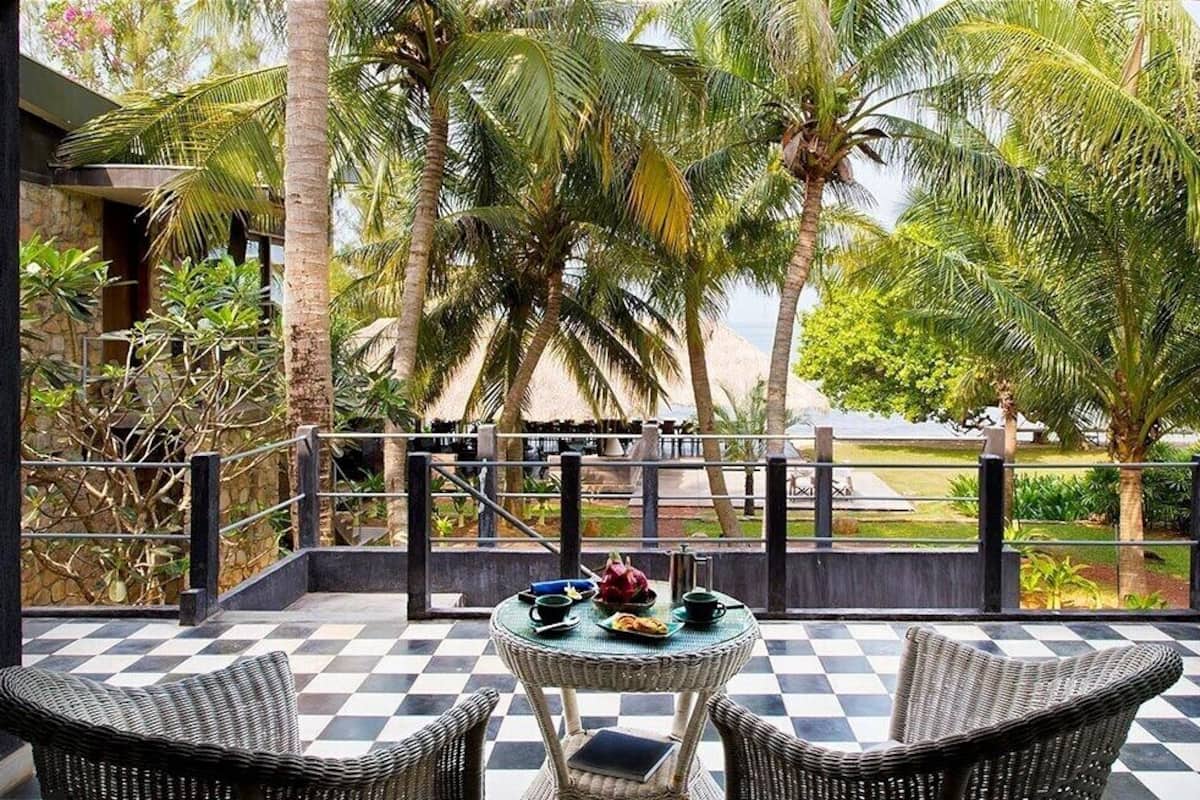 A checkered outdoor seating area with wicker furniture, a table with a cup and saucer, and a view of palm trees and a building in the background.