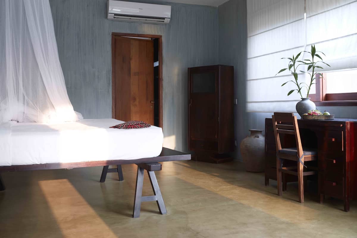 A bedroom with a bed, a wooden wardrobe, a desk with a chair, and a potted plant.